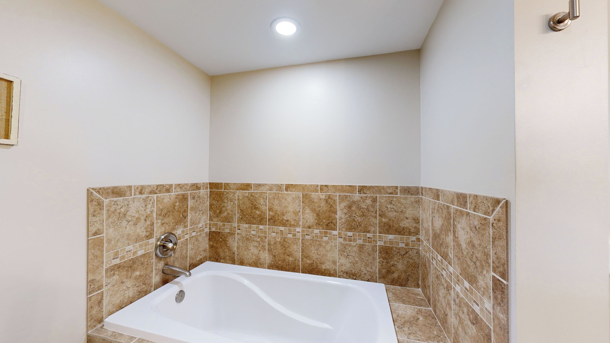 A bathroom with a white bathtub, beige tiled walls, and a recessed ceiling light.
