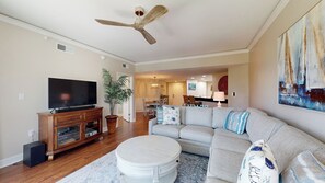 Un salon comprenant un canapé d’angle, un téléviseur, un meuble de rangement en bois, un ventilateur de plafond et, au fond, un coin repas.