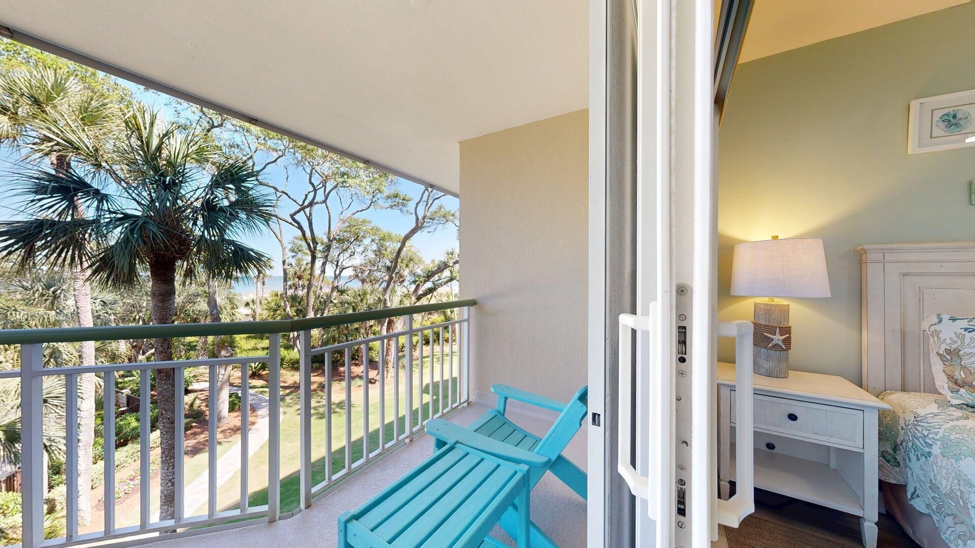 A balcony with a view of palm trees and a beach, a white nightstand with a lamp, and a bed with a floral bedspread.