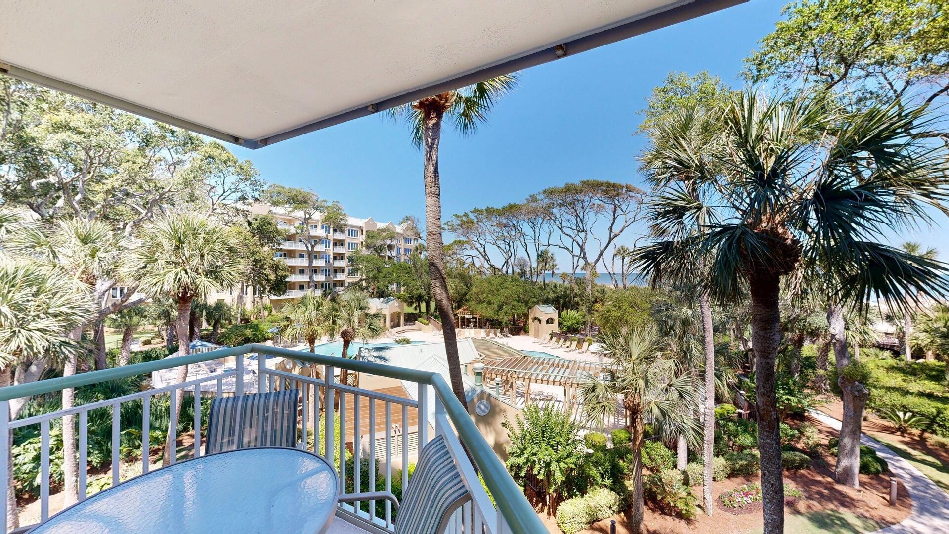 A balcony with a glass table and chairs, overlooking a pool area with lounge chairs and palm trees.