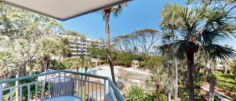 A balcony with a glass table and chairs, overlooking a pool area with lounge chairs and palm trees.