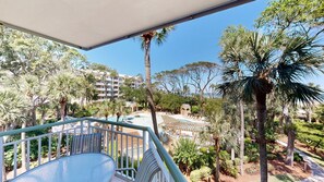 A balcony with a glass table and chairs, overlooking a pool area with lounge chairs and palm trees. - Oceanfront Coastal One Bedroom Corner Villa! (Hilton Head)