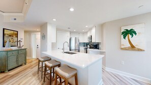 A modern kitchen with a central island, bar stools, and a painting of a palm tree. - Oceanfront Coastal One Bedroom Corner Villa! (Hilton Head)
