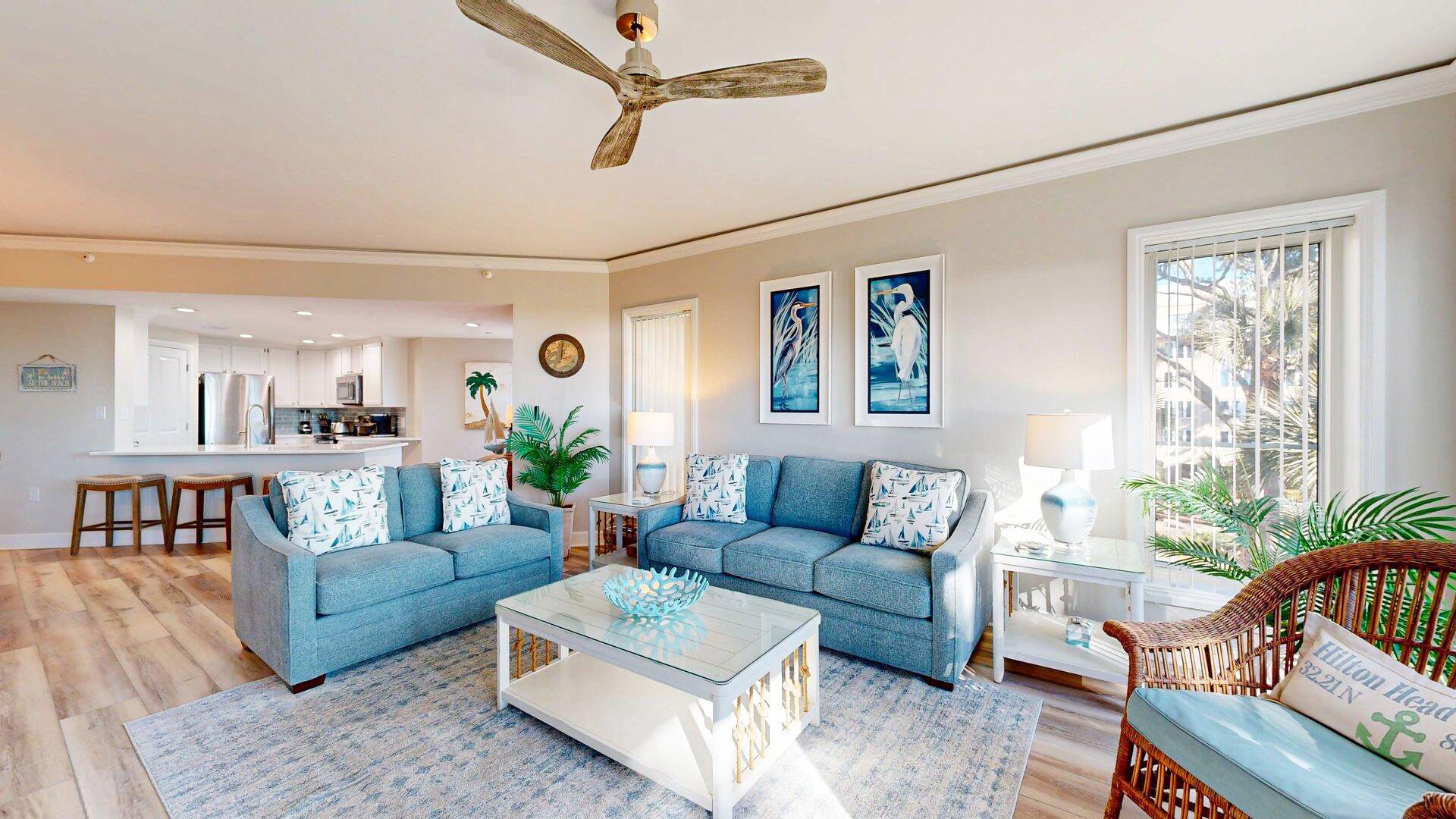 A spacious living area with a modern kitchen in the background, a large window with a view, and a ceiling fan.