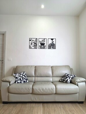 A neutral-toned leather sofa with two patterned throw pillows, a wall-mounted light fixture, and three framed abstract wall art pieces.