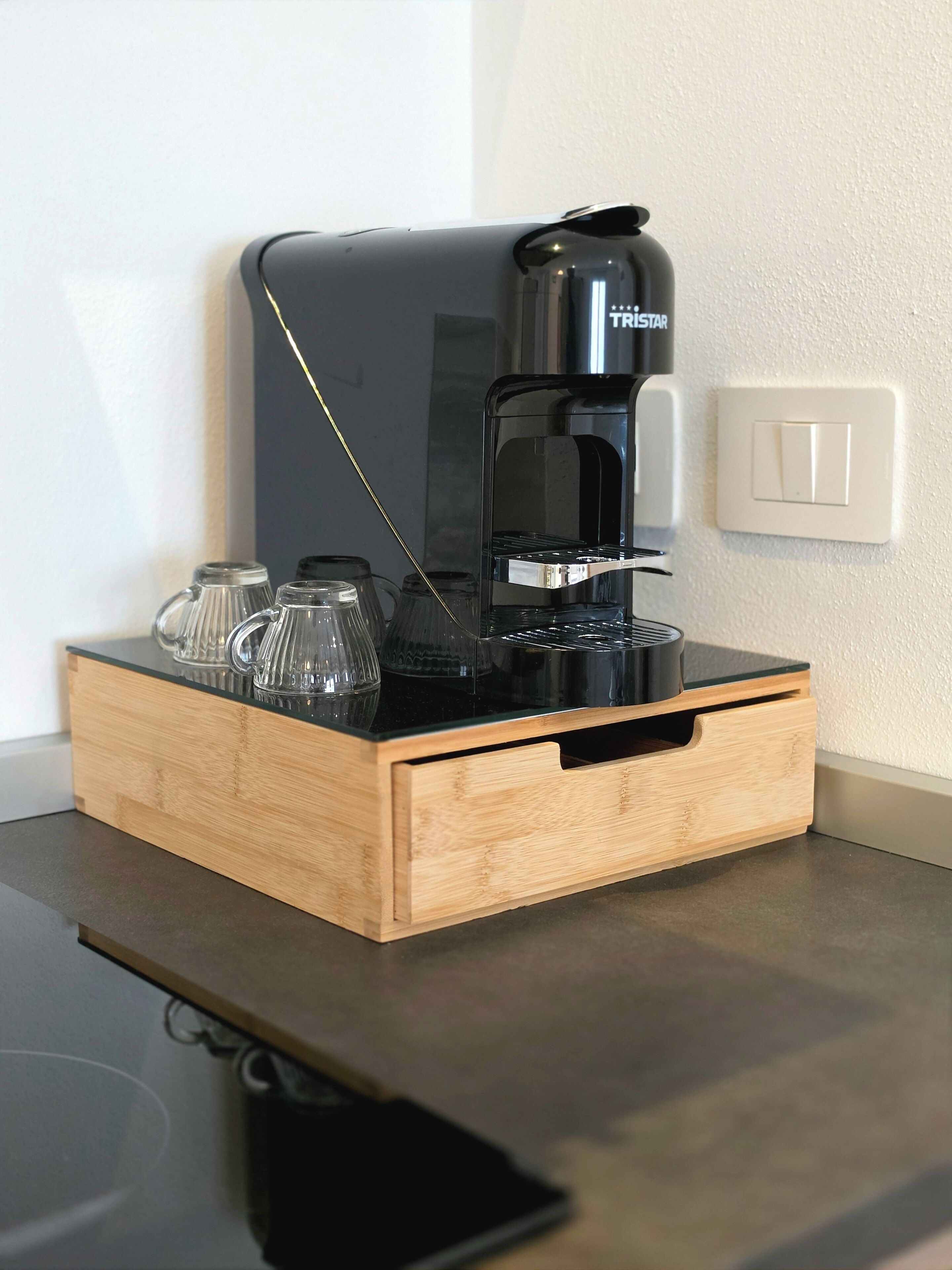 Een koffiezetapparaat op een houten dienblad met een lade eronder, naast een stopcontact.