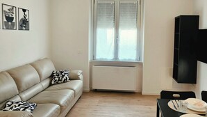 Un salon avec un canapé beige, une table à manger avec des chaises, une fenêtre avec des stores et un meuble TV noir.
