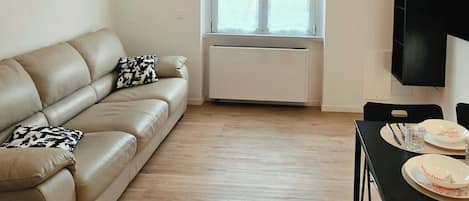 Una sala de estar con un sofá beige, una mesa de comedor con sillas, una ventana con persianas y un mueble de TV negro.
