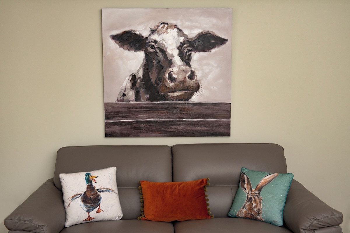 A living room with a brown leather sofa, a wall-mounted painting of a cow, and three decorative pillows with animal prints.
