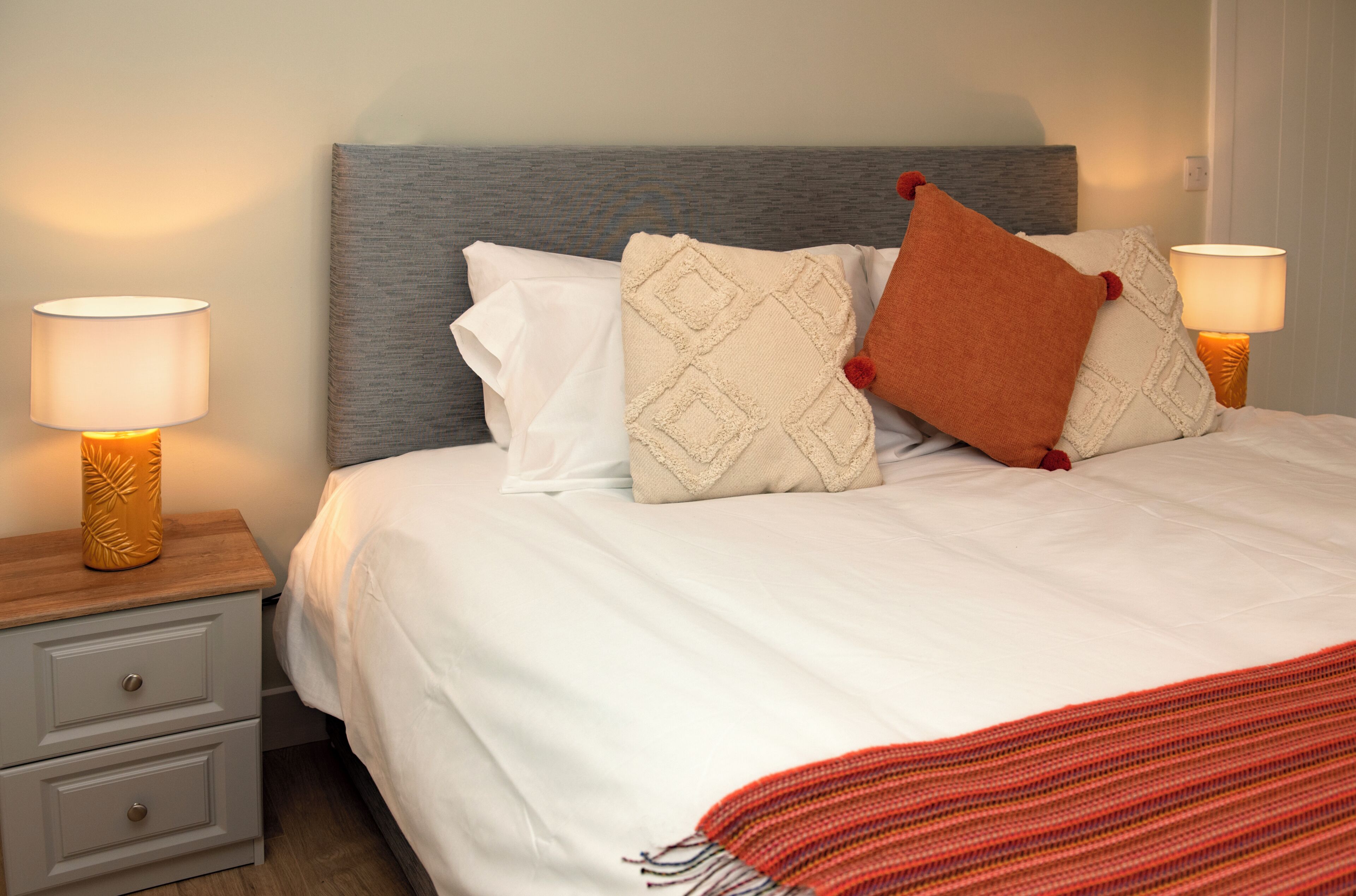 A neatly made bed with a grey headboard, white bedding, and decorative pillows. There are two bedside tables with lamps, one on each side of the bed.
