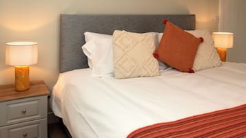 A neatly made bed with a grey headboard, white bedding, and decorative pillows. There are two bedside tables with lamps, one on each side of the bed.