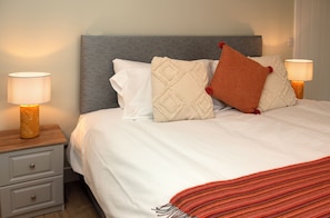 A neatly made bed with a grey headboard, white bedding, and decorative pillows. There are two bedside tables with lamps, one on each side of the bed.