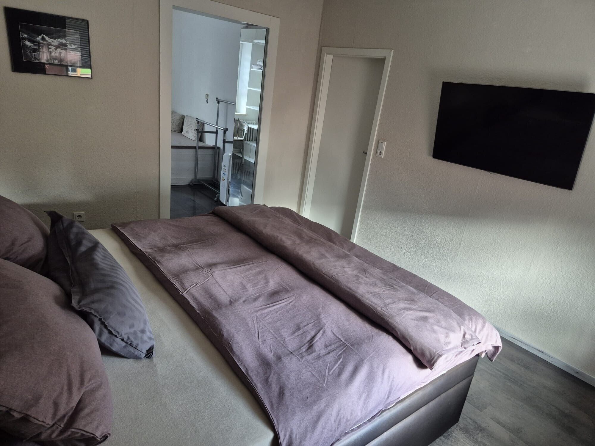 Ein Zimmer mit einem Bett, einem flachbildfernseher an der Wand und einer Tür zu einem weiteren Raum.