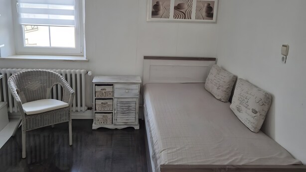 A bedroom with a bed, a chair, a nightstand, a radiator, and a window with blinds. - Guesthouse 'Eifelglück' with Private Terrace, Garden, and Wi-Fi (Großlittgen)