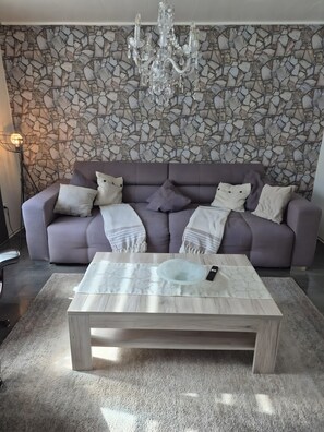 A living room with a stone wall, a grey sofa, a coffee table, and a chandelier. - Guesthouse 'Eifelglück' with Private Terrace, Garden, and Wi-Fi (Großlittgen)