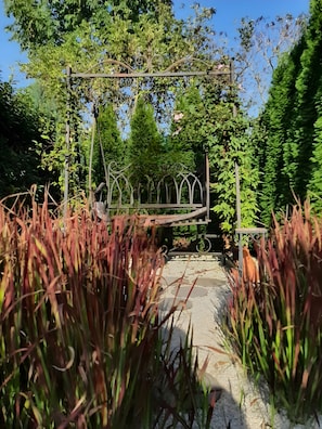 Een tuinpad met een metalen boog, omringd door weelderige begroeiing en hoge grassen.