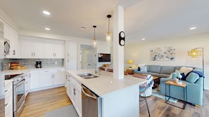 A modern kitchen with white cabinetry, a central island, and a dining area with a grey sofa and blue cushions. - Beautiful Three Bedroom Villa with Sweeping Views of George Fazio Golf Course (Hilton Head)