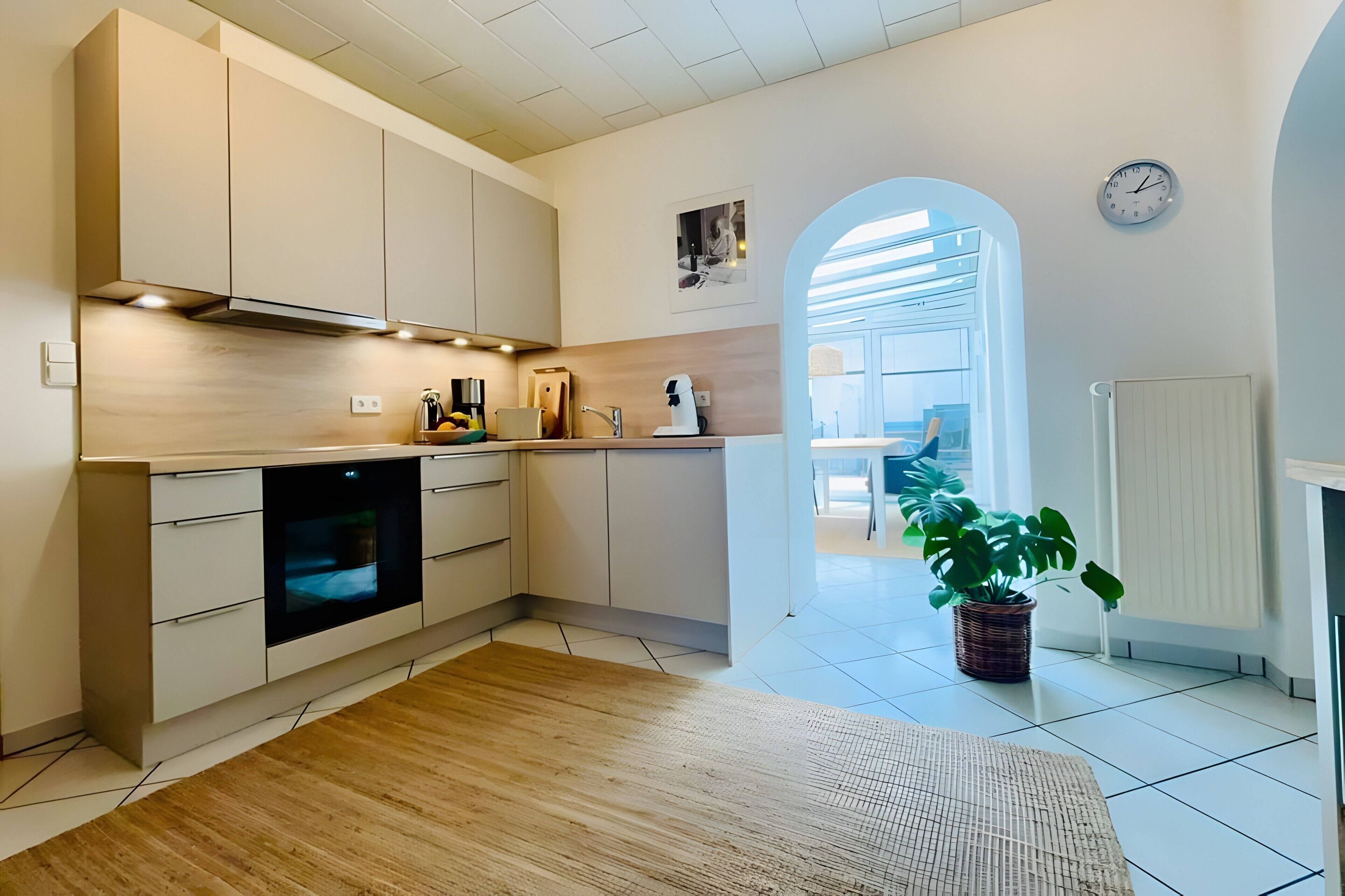 A modern kitchen with a built-in oven, white cabinets, and a wooden floor. There is a potted plant and a clock on the wall.