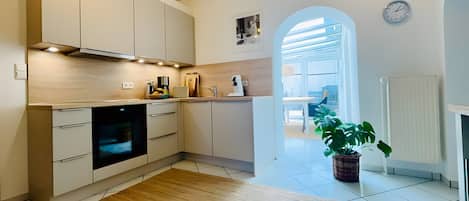 Una cocina moderna con horno empotrado, armarios blancos y suelo de madera. Hay una planta en maceta y un reloj en la pared.