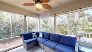 Un espacio de descanso techado al aire libre con un sofá modular azul, un ventilador de techo y grandes ventanales que ofrecen vistas de los árboles circundantes.