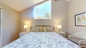 A bedroom with a bed, two bedside tables with lamps, a window with a view of trees, and a wicker chair. - 4 Bedroom 4 Bath house on the 11 mile lagoon (Hilton Head)