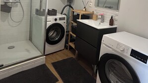 A compact bathroom with a glass shower enclosure, a washing machine, a dryer, and a sink with a mirror above it. - LE REFUGE DU MOULIN - 2-Bedroom Apartment in Downtown Senlis (Parking) (Senlis)