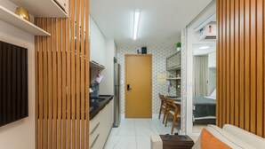 Uma cozinha compacta com geladeira, pia e painéis de madeira. É visível uma área de jantar com cadeiras e mesa, e uma porta dá acesso a um quarto com cama e cortinas.