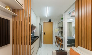 Eine kleine Küche mit Kühlschrank, Spüle und Holzvertäfelung. Ein Essbereich mit Stühlen und Tisch ist zu sehen, und eine Tür führt zu einem Schlafzimmer mit Bett und Vorhängen.