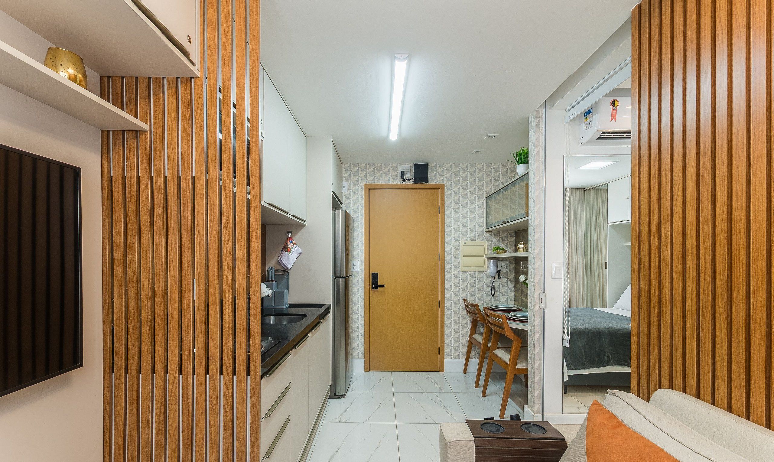 A compact kitchen with a refrigerator, sink, and wooden paneling. A dining area with chairs and a table is visible, and a door leads to a bedroom with a bed and curtains.