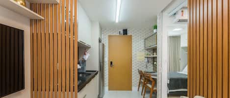 A compact kitchen with a refrigerator, sink, and wooden paneling. A dining area with chairs and a table is visible, and a door leads to a bedroom with a bed and curtains.