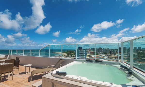 Een dakterras met een hottub, rieten meubels en een glazen balustrade met uitzicht op een stadsgezicht en de oceaan.