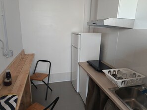 A compact kitchen with a wooden bench, a chair, a white refrigerator, a stove, and a sink with a dish rack.