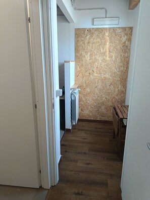 A narrow hallway with wooden flooring, a white door on the left, and a kitchen area with a wooden wall and a radiator on the right.