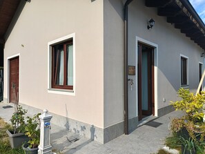 A building with a beige facade, two windows with wooden frames, a door, and a small garden area with potted plants.