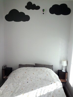 Un dormitorio con cama, almohadas, mesita de noche con lámpara y decoración en la pared que incluye nubes y un globo aerostático.