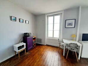 Een kleine, goed verlichte kamer met een houten vloer, een wit bureau, een stoel, een paars kastje en een raam met jaloezieën.