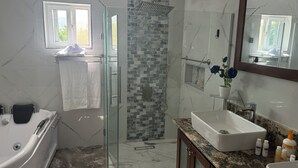 Uma casa de banho moderna com chuveiro em cabine de vidro, soalho em mosaico de mármore, banheira branca e bancada de granito com lavatório e produtos de higiene pessoal.