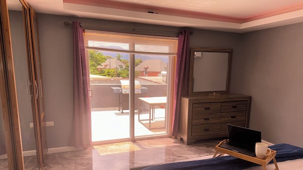 Quarto com cama, cômoda, laptop sobre um suporte e porta de vidro deslizante que dá para um pátio externo.