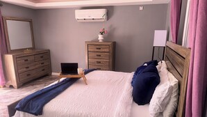 Quarto com cama, cômoda de madeira, escrivaninha com laptop, um vaso de flores e um ar-condicionado na parede.