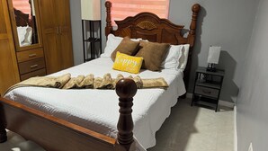 Uma cama bem arrumada com uma almofada rotulada 'my happy place', um abajur e um guarda-roupa de madeira.