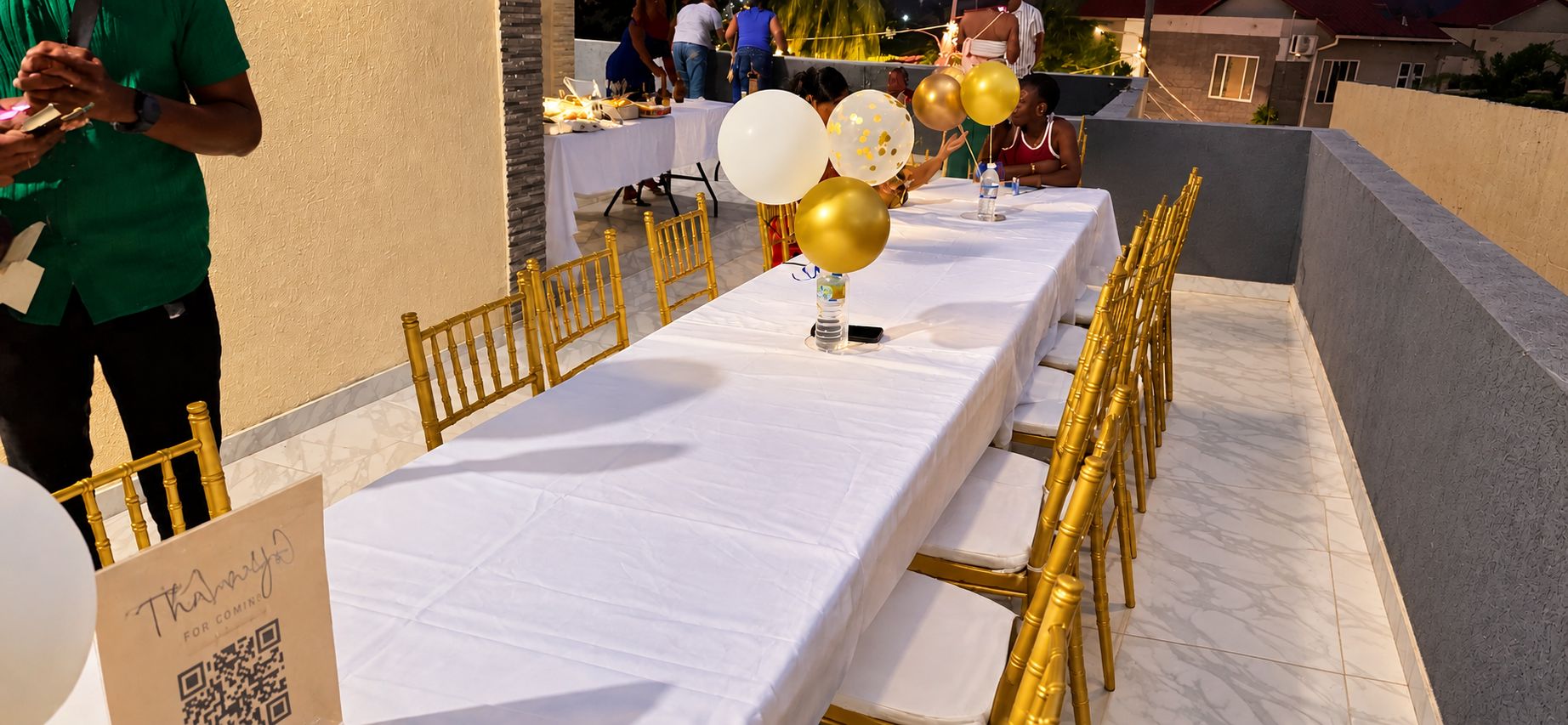A long table with white tablecloths and gold chairs, set up outdoors with balloons and a QR code sign.