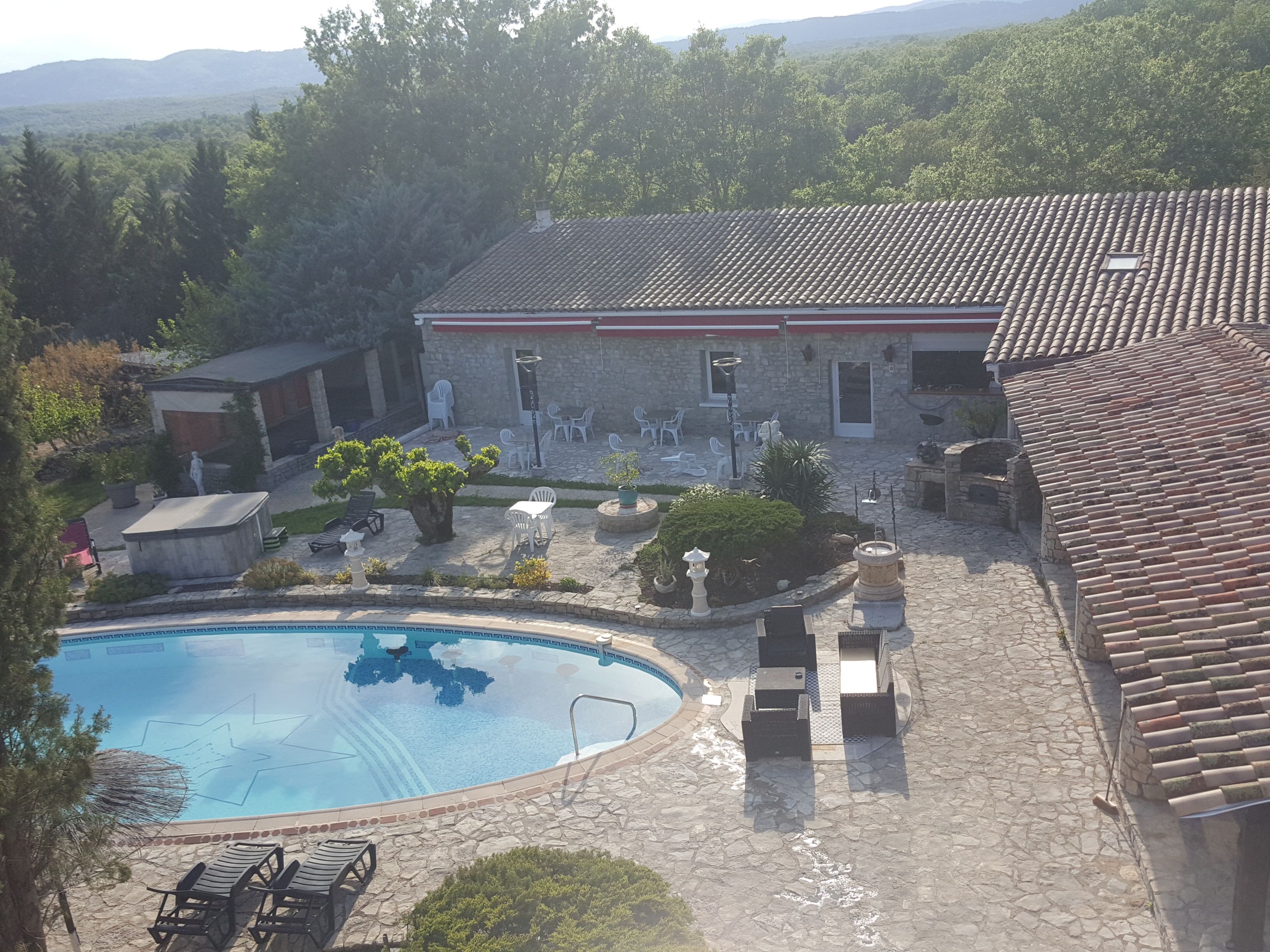 Un espace piscine avec des chaises longues, un bâtiment en pierre doté d’un toit en tuiles et une végétation luxuriante tout autour.
