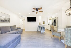A modern living room with a grey sofa, a ceiling fan, a flat-screen TV, and a kitchen area with a fridge and bar stools. - Baby Beach 2BR, Walk to Everything W/Parking & AC! (Newport Beach)