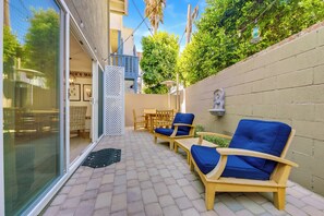 Un patio exterior con sillas y mesa de madera, puerta de cristal y una lámpara fijada a la pared.
