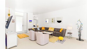 Uma sala de estar moderna com um sofá, pufes, uma mesa de centro, uma televisão e um abajur.