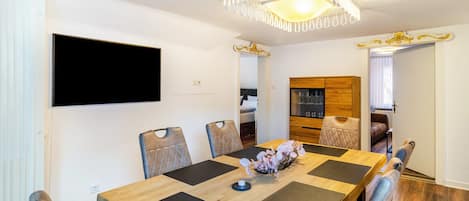 A dining area with a wooden table, cushioned chairs, a chandelier, and a television mounted on the wall.