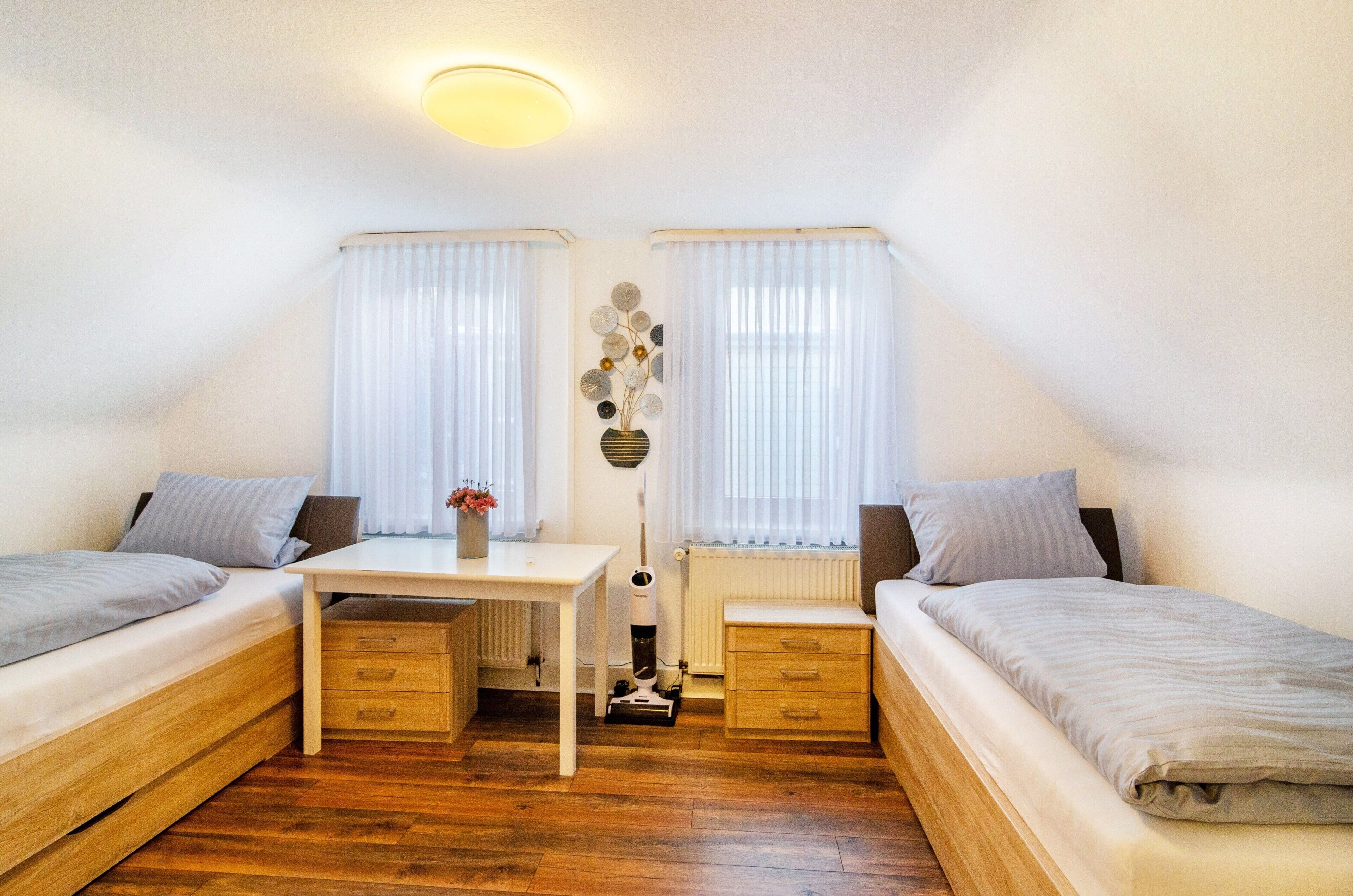Ett litet, väl upplyst rum med två sängar, ett skrivbord, ett element och en dekorativ vas.