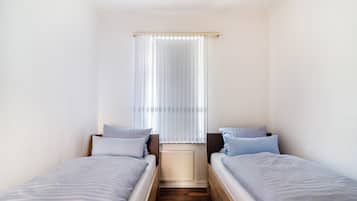 Un petit logement composé d’une seule pièce, avec deux lits simples, une fenêtre avec des rideaux légers et un radiateur.