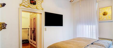 A bedroom with a bed, a TV mounted on the wall, a ceiling fan, and a door leading to another room.
