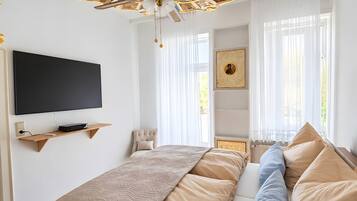 Ein Schlafzimmer mit einem großen Bett, einem Deckenventilator, einem an der Wand montierten Fernseher und einem Fenster mit durchscheinenden Vorhängen.